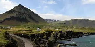 Road in Iceland leading up to a white house overlooking the sea with mountain in the background